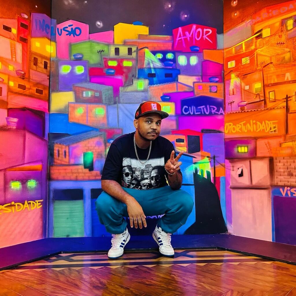 Foto do artista Load, sagachado em frente a uma pintura de casas periféricas